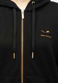 Sweatshirt preto com capuz e fecho dourado, apresentando logótipo bordado e cordões. Material suave, bolsos frontais e design sem costuras.