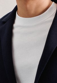 Vit rundhalsad t-shirt som bärs under en mörkblå kavaj. Kavajen har en texturerad tyg och ficklappar. Närbild på halsringningen.