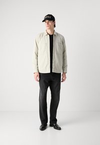 Light beige padded jacket with two chest pockets, worn over a black shirt, paired with dark jeans and black shoes, and a black cap marked "BOSS."