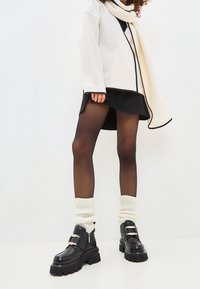 Bottes noires à la cheville avec des semelles épaisses, associées à des chaussettes blanches à côtes jusqu'aux genoux. La tenue comprend un haut crème avec des bordures noires et une jupe noire.