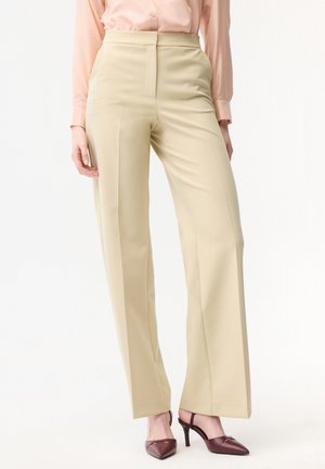 Frau trägt beige, hoch taillierte, weit geschnittene Hosen, ein hellrosa Hemd mit Knöpfen und burgunderrote spitze Absätze mit Riemchen.