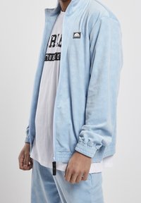 Světlá modrá bunda na zip s texturovanou látkou, žebrovanými manžetami a malou nášivkou s logem. Nošena přes bílé tričko s potiskem.