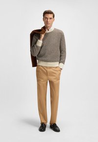 Suéter de punto en marrón y crema con patrón texturizado, combinado con pantalones color beige. Zapatos oscuros completan el conjunto. Chaqueta marrón sobre el hombro.