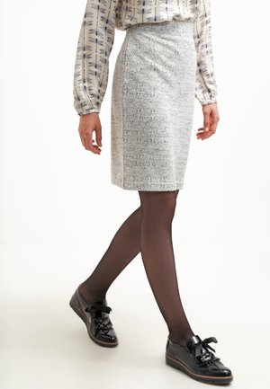 Kobieta w szarej ołówkowej spódnicy o fakturze, przezroczystych czarnych rajstopach, czarnych lakierowanych mokasynach z kokardkami oraz bluzce z wzorem ważek.