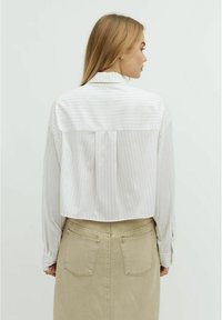 Camicia bianca con strisce verticali rosa, maniche lunghe e orlo corto, abbinata a una gonna beige con tasche posteriori.