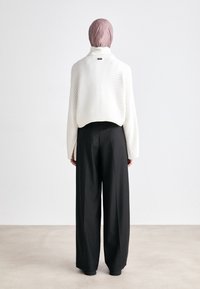 Weißer Rippstrickpullover mit hohem Kragen, kombiniert mit schwarzen, hoch taillierten, weiten Hosen. Das Outfit wird mit einem pastellrosa Kopftuch abgerundet.