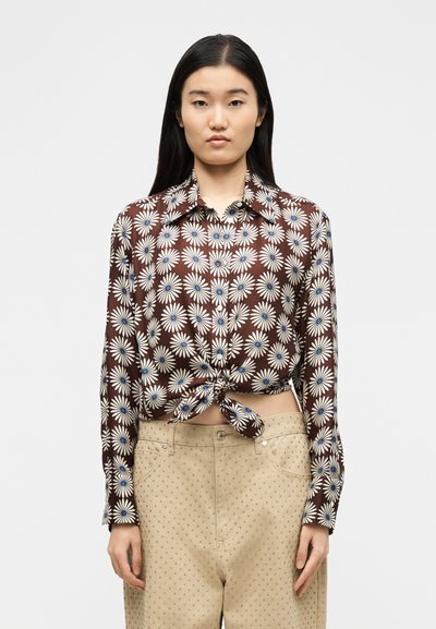 Νεαρή γυναίκα με μακριά ίσια σκούρα μαλλιά, ντυμένη με καφέ φλοράλ μπλούζα δεμένη στο πλάι και μπεζ ψηλόμεσα παντελόνια διακοσμημένα με τρουκς, στέκεται μπροστά σε απλό φόντο.