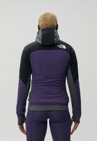 Lilla og svart zip-up jakke med hette, som har en tettsittende design, teksturert stoff og logo på baksiden. Lange ermer med tommelhull.