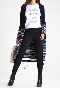 Granatowy i kolorowy kardigan w paski, biały t-shirt z nadrukiem, czarne legginsy i czarne botki. T-shirt zawiera cytat w czarnym tekście.