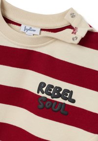 Rood en crème gestreepte pullover met ronde hals, met een patch waarop "REBEL SOUL" in het zwart staat en drukknoopjes op de schouder.
