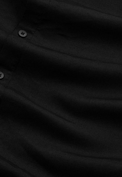 Una maglietta nera in tessuto liscio presenta una chiusura frontale con due bottoni visibili e leggere variazioni di texture sulla superficie.