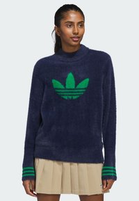 Marineblå fuzzy genser med grønn Adidas-logo og striper på ermer. Høy hals og avslappet passform, laget av mykt, strukturert materiale.