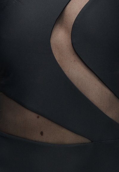 Gros plan sur un tissu noir avec une découpe en filet transparent laissant apparaître la peau avec deux petits grains de beauté en dessous.