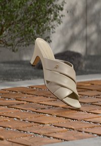 Μπεζ σανδάλι με ψηλό τακούνι και σταυρωτούς ιμάντες, με μικρό χρυσό λογότυπο, πάνω σε ηλιαζόμενη ξύλινη επιφάνεια πλακιδίων σε εξωτερικό χώρο.