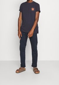 T-shirt navy a maniche corte con un design circolare sul petto, abbinato a pantaloni scuri e sandali marroni, evidenziando uno stile casual.