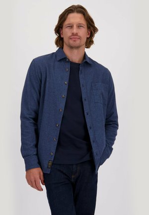 Man met schouderlang haar in een blauwe overhemden met knopen over een marineblauwe trui en donkere jeans, die staat met één hand in zijn zak.