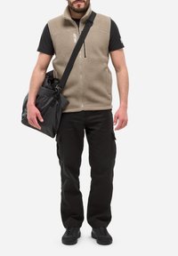 Mann trägt eine beigefarbene Fleece-Weste, ein schwarzes T-Shirt, schwarze Cargo-Hosen und schwarze Sneaker, und trägt eine schwarze Sporttasche mit einem Schultergurt.