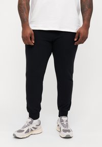 Svart joggebukse med en smal design, ribbede mansjetter og en myk tekstur, kombinert med lysegrå og kremfargede sneakers.