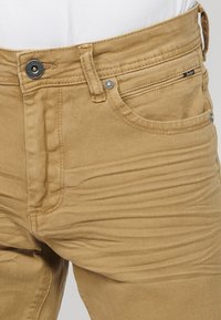 Jean en denim beige avec une coupe droite, présentant des coutures visibles, une poche avant et un bouton de fermeture en chrome. Texture lisse, design décontracté.