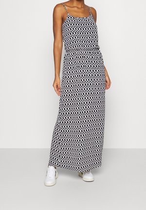 Vrouw draagt een maxi-jurk met een zwart-wit geometrisch patroon, dunne bandjes en witte sneakers, staand tegen een effen achtergrond.