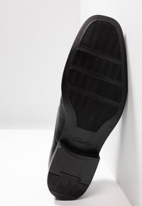Zwarte lederen schoen met een gestructureerde rubberen zool, voorzien van horizontale groeven en een opvallend Clarks-logo op de hiel.
