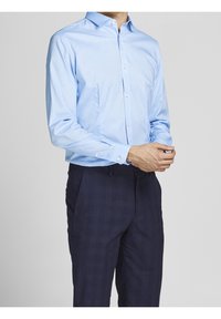 Person som bär en ljusblå långärmad skjorta med knappar och mörkblå rutiga byxor, stående med händerna hopknäppta.