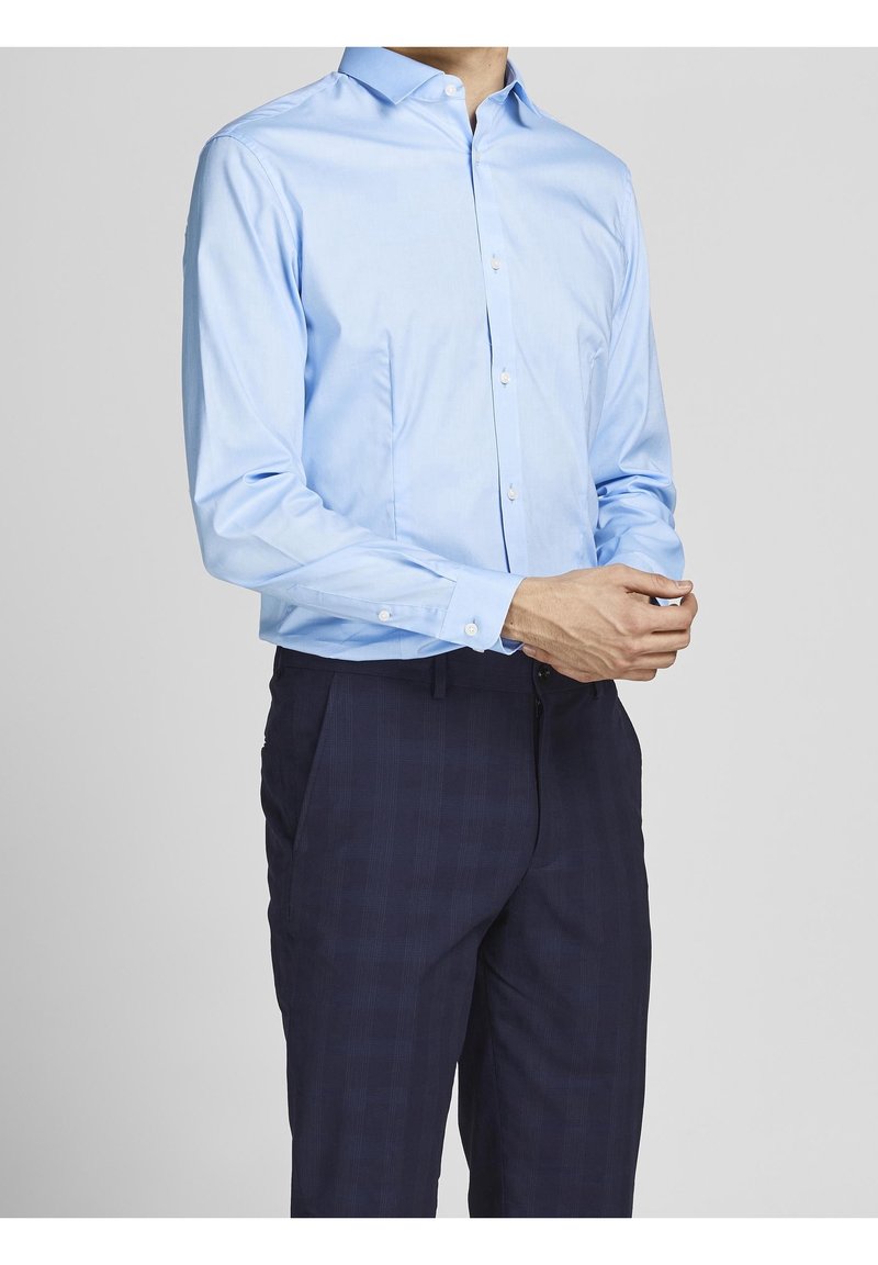 Person som bär en ljusblå långärmad skjorta med knappar och mörkblå rutiga byxor, stående med händerna hopknäppta.