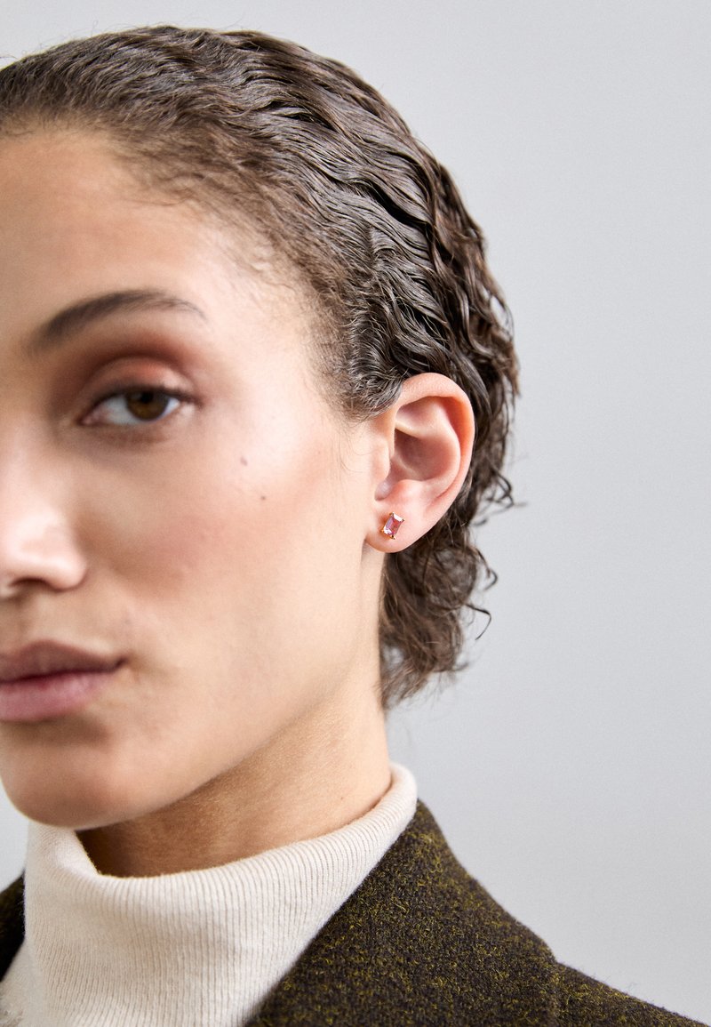 Rožu zelta auskars ar taisnstūra formas dārgakmeni, novietots uz strukturēta fona, kopā ar gaismas krāsas augstu kaklu un tumšu uzvalku.