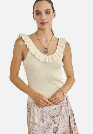 Mujer con blusa sin mangas color crema con volantes, collar dorado y falda con estampado floral, manos ligeramente entrelazadas a la altura de la cintura.