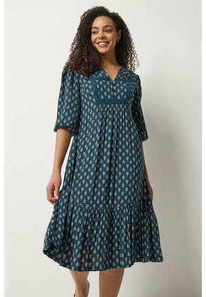 Femme souriante aux cheveux bouclés portant une robe turquoise longueur genou avec des motifs floraux blancs, des manches bouffantes et un ourlet froncé, debout contre un mur uni.