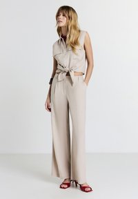 Donna in piedi che indossa un top corto senza maniche beige legato e pantaloni larghi, con sandali con tacco aperti rossi, con le mani nelle tasche.
