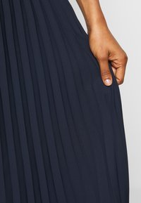 Tessuto di abito a pieghe blu navy con una consistenza liscia, caratterizzato da pieghe orizzontali e una mano che afferra il materiale vicino all'orlo.