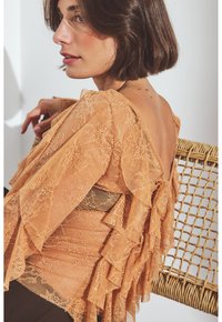 Woman with short brown hair wearing a peach lace blouse with ruffles sitting sideways on a woven chair against a white background.