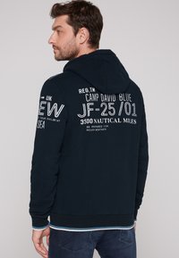Felpa navy con grafiche bianche sul retro, dotata di tasca a marsupio e polsini a coste. Tessuto morbido, vestibilità casual.