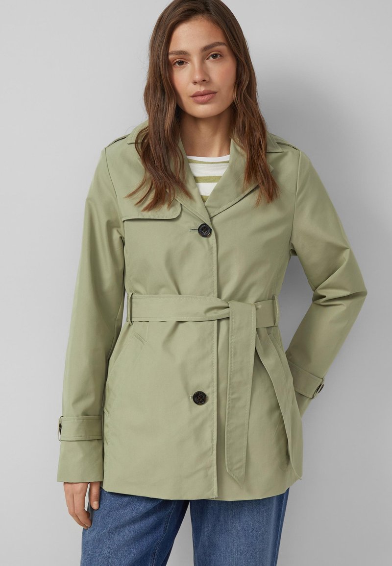 Frau trägt einen hellgrünen Trenchcoat mit Gürtel über einem gestreiften Hemd und blauen Jeans, steht vor einem schlichten grauen Hintergrund.