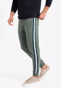 Grüne Sweatpants mit seitlichen Streifen in Marineblau und Weiß. Gerippte Bündchen, Fronttasche und eine lockere Passform. Getragen mit hellen Schuhen.