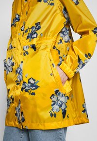 Chaqueta amarilla impermeable con patrón floral azul, que cuenta con un cierre frontal, bolsillos y puños elásticos. Acabado de satén suave.