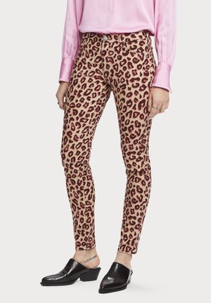 Femme portant un pantalon skinny beige et marron à imprimé léopard, un chemisier en satin rose et des mules à talons en cuir noir, debout devant un fond uni.