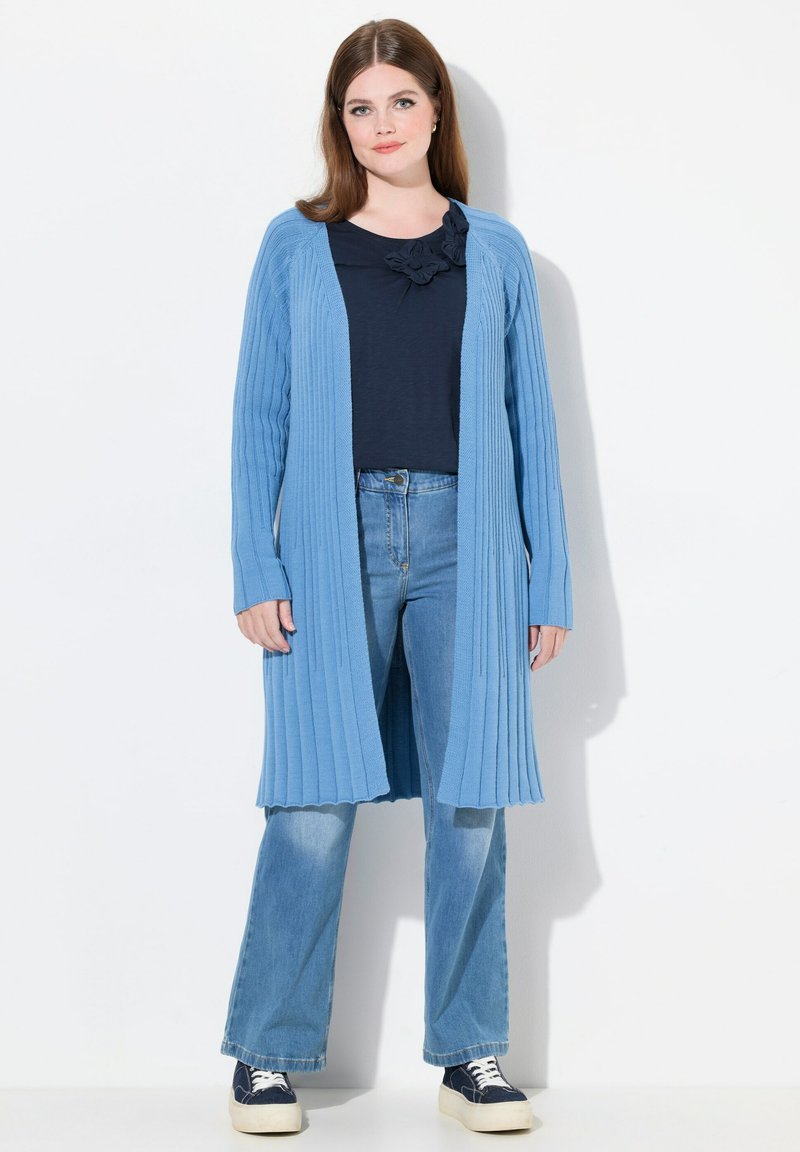 Blaues geripptes Cardigan mit langen Ärmeln, offener Vorderseite und strukturiertem Stoff, über einem dunklen Oberteil getragen und kombiniert mit hellblauen Jeans.