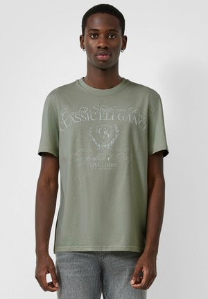 Junger Mann, der ein olivgrünes T-Shirt mit Grafik und graue Jeans trägt, steht vor einem schlichten hellgrauen Hintergrund.