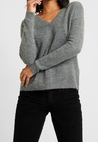 Pull en maille grise avec col en V, manches longues et coupe légèrement ample. Associates à un pantalon en velours côtelé noir, présentant un motif texturé.
