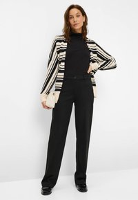 Cardigan rayé noir et blanc sur un top noir, associé à un pantalon noir taille haute et des chaussures noires. Tenant un sac à main crème.