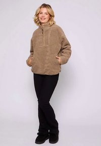 Beżowy polarowy pulower z półzamkiem przy kołnierzu, długimi rękawami i przednimi kieszeniami, w połączeniu z czarnymi spodniami i czarnym obuwiem.