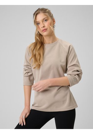 Młoda kobieta z długimi blond włosami w beżowej bluzie i czarnych spodniach pozująca na szarym tle.