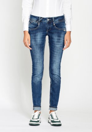 Personne portant un jean skinny bleu retroussé, une chemise blanche à manches longues et des baskets blanches avec des accents verts, debout devant un fond uni.