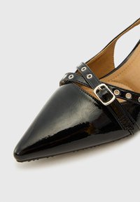 Musta patentoitu nahkainen slingback-kengät teräväkärkisinä, koristeellinen hihna, jossa on pyöreitä aukkoja, ja hopea solkiyksityiskohta.