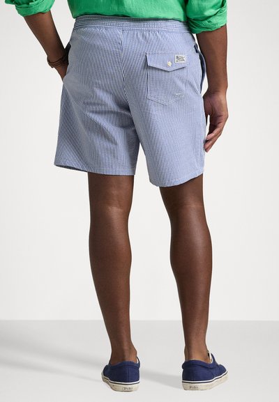 Shorts rayés bleu et blanc pour homme avec une poche arrière, fabriqués en tissu léger, dotés d'une ceinture élastique. Portés avec des chaussures bleues.