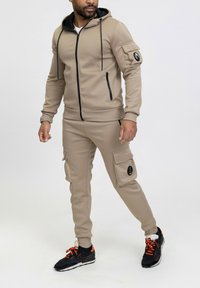 Haut de survêtement beige avec fermeture éclair noire et cordons, pantalon assorti avec poches cargo et baskets noires avec accents orange. Texture lisse.