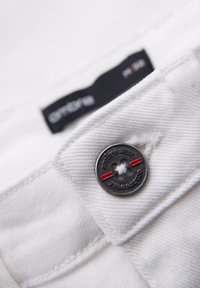 Tejido blanco con textura visible, con un botón plateado con acentos rojos y texto grabado. Etiqueta negra parcialmente visible en el fondo.