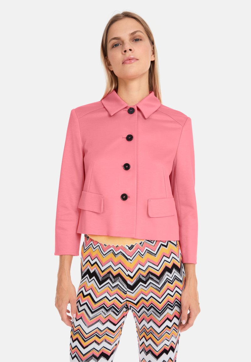Veste courte rose avec un col, ornée de boutons noirs et de deux poches avant. Associée à un pantalon en chevrons multicolores.